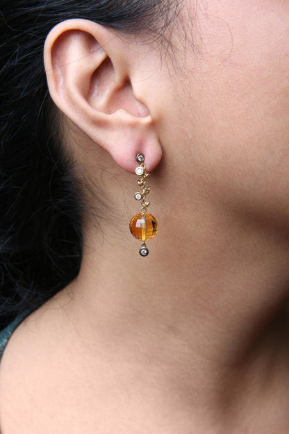 Close-up side view of a model wearing yellow metal drop earrings featuring a large faceted yellow gemstone bead and multiple small round bezel-set white and brown gemstones arranged along the chain.