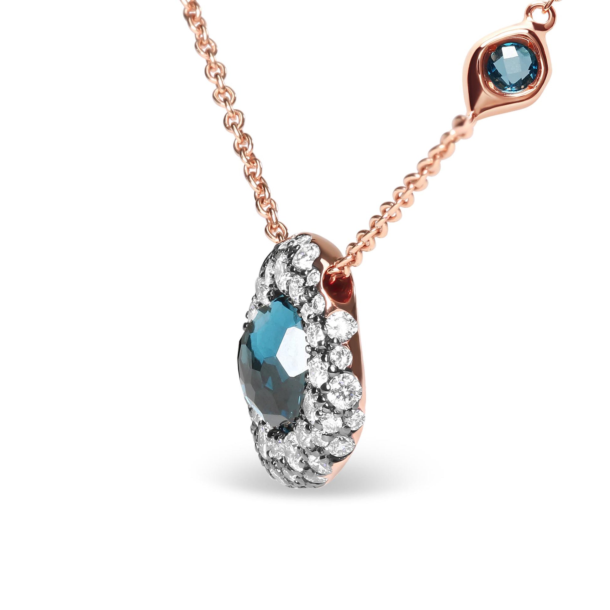 Close-up front angled view of a rose metal necklace with a pendant featuring a central blue gemstone surrounded by multiple prong-set white gemstones, and a small bezel-set white gemstone and a small bezel-set blue gemstone on the chain.