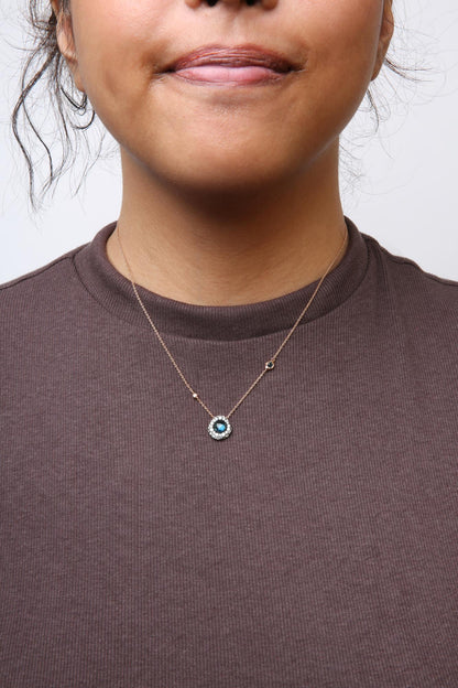 Front view of a rose metal necklace with a round blue gemstone surrounded by white gemstones, worn on a model.