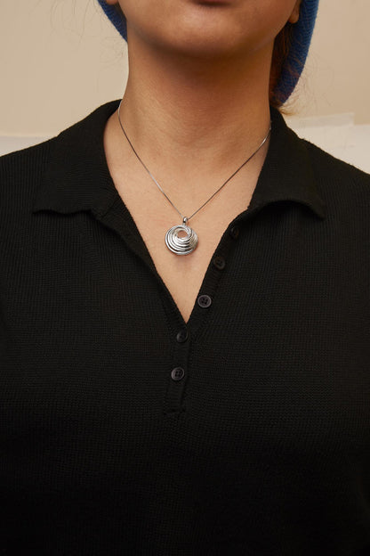 Close-up front view of a white metal necklace with a circular, layered pendant worn on a model with a black buttoned top and blue headwear.