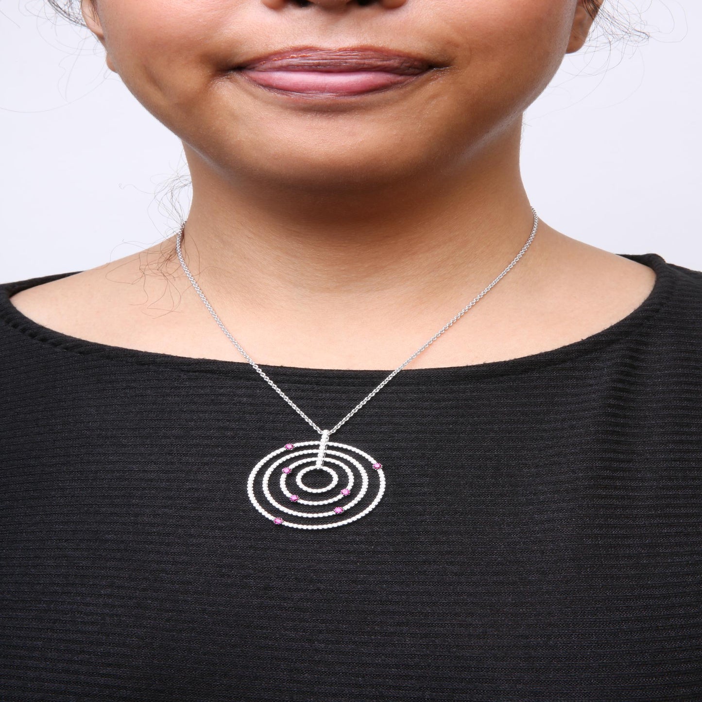 Close-up front view of a woman's neck wearing a white metal necklace with a circular pendant featuring multiple concentric rings adorned with white gemstones and red gemstones, shown against a black top.