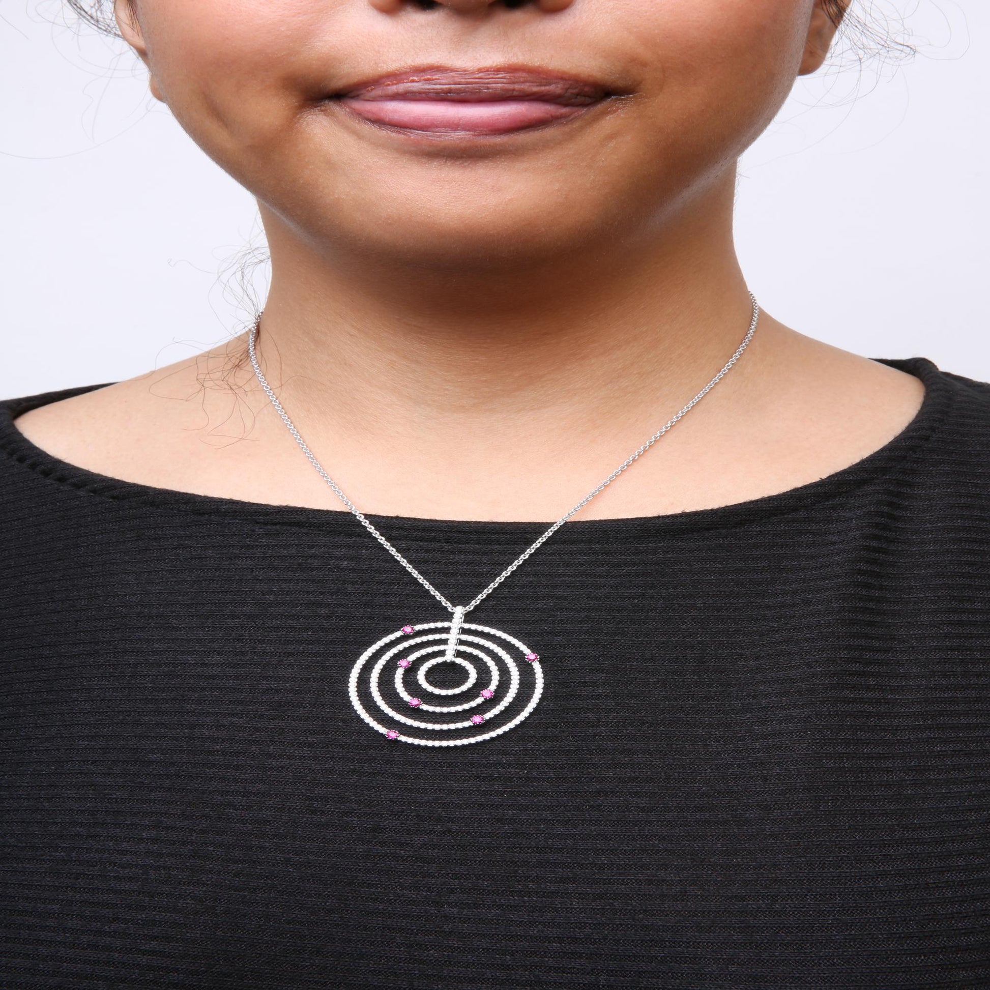 Close-up front view of a woman's neck wearing a white metal necklace with a circular pendant featuring multiple concentric rings adorned with white gemstones and red gemstones, shown against a black top.
