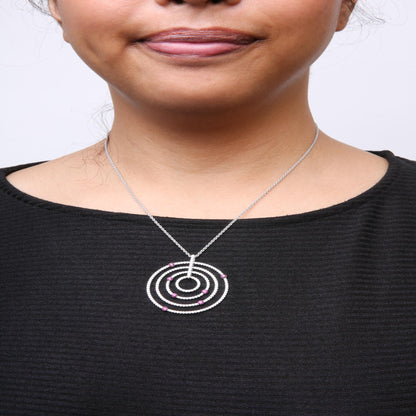 Close-up front view of a woman's neck wearing a white metal necklace with a circular pendant featuring multiple concentric rings adorned with white gemstones and red gemstones, shown against a black top.