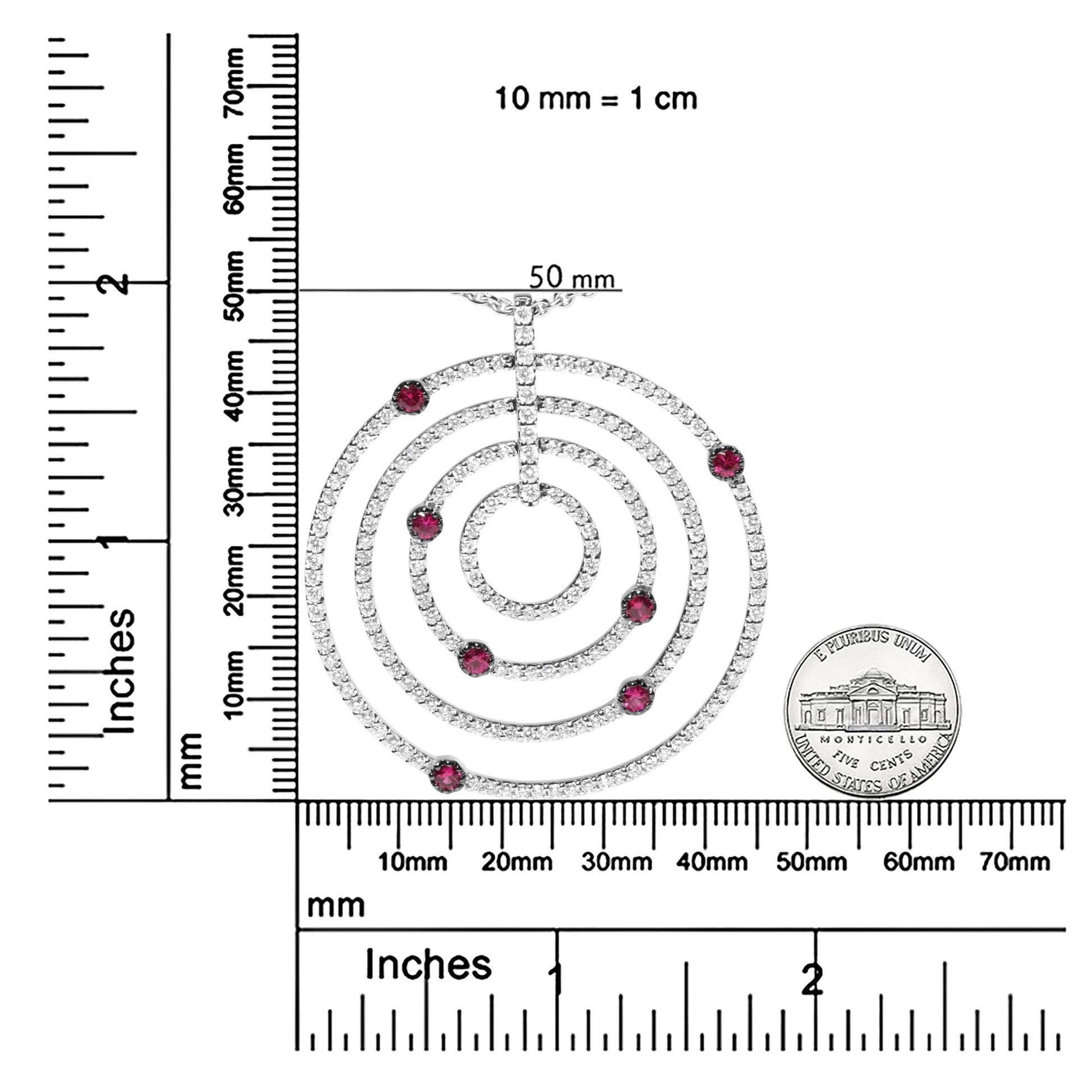 Top-down view of a white circular necklace pendant with concentric rings encrusted with small white gemstones and accented with round red gemstones, shown on a ruler and scale for size reference.