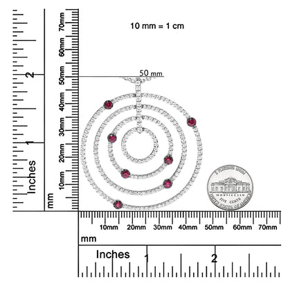 Top-down view of a white circular necklace pendant with concentric rings encrusted with small white gemstones and accented with round red gemstones, shown on a ruler and scale for size reference.