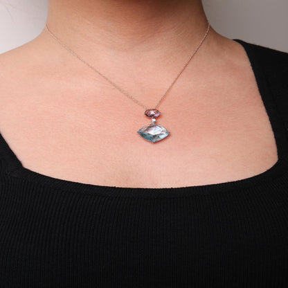 Rose metal necklace worn on model, featuring a blue cushion-shaped gemstone pendant and a smaller round pink gemstone above it, separated by a small white round gemstone; shot from front.