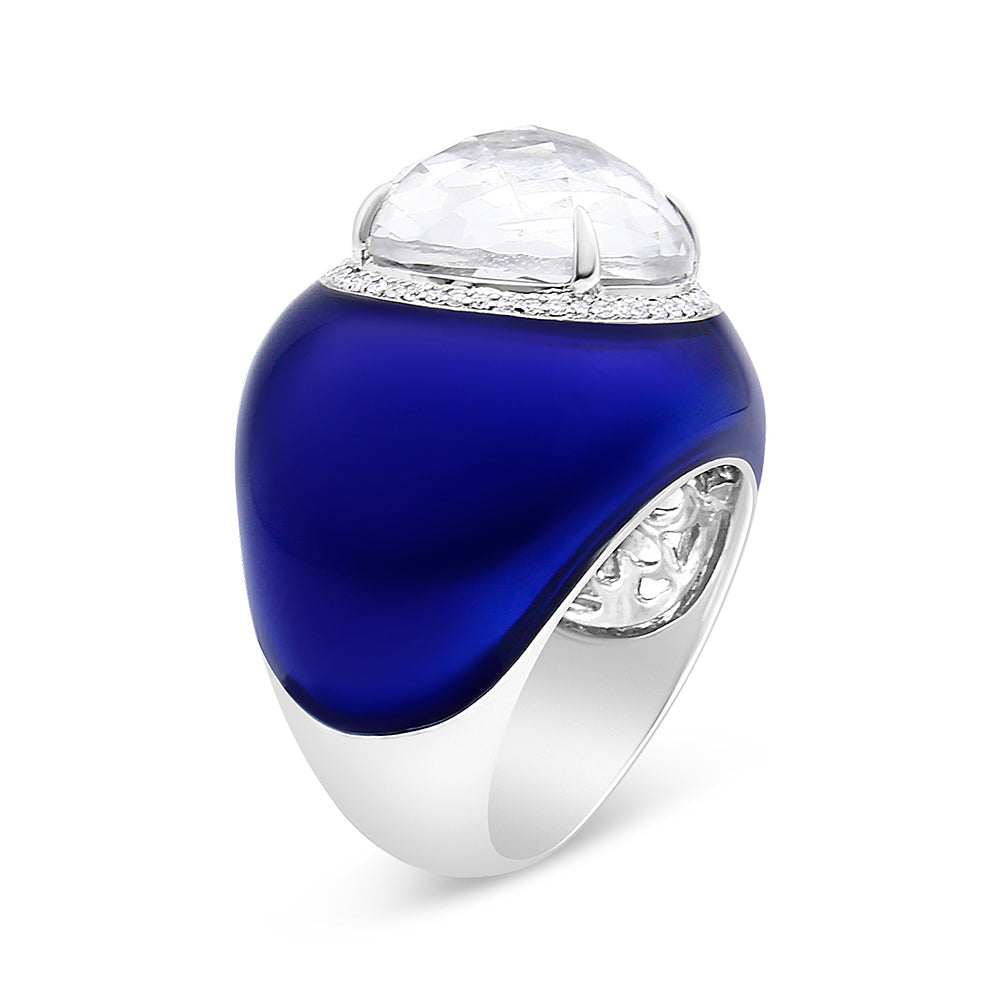 White metal ring with a smooth curved blue enamel surface, topped with a large round white gemstone in bezel setting surrounded by a row of small round white gemstones in shared prong setting, shown at an angle.
