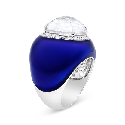 White metal ring with a smooth curved blue enamel surface, topped with a large round white gemstone in bezel setting surrounded by a row of small round white gemstones in shared prong setting, shown at an angle.