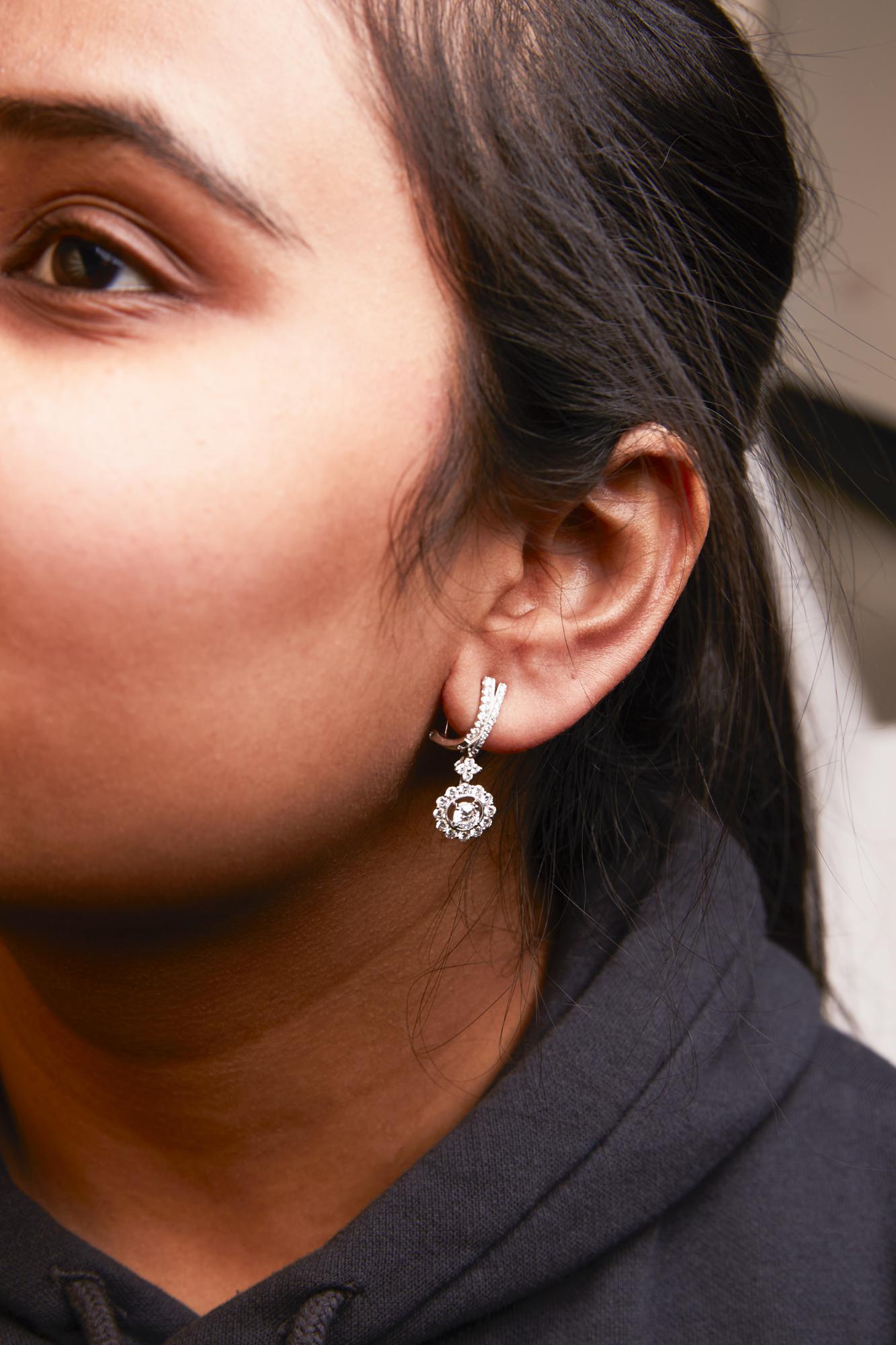 Close-up side view of a woman's ear wearing white metal drop earrings with two larger round prong-set white gemstones surrounded by smaller round prong-set white gemstones.