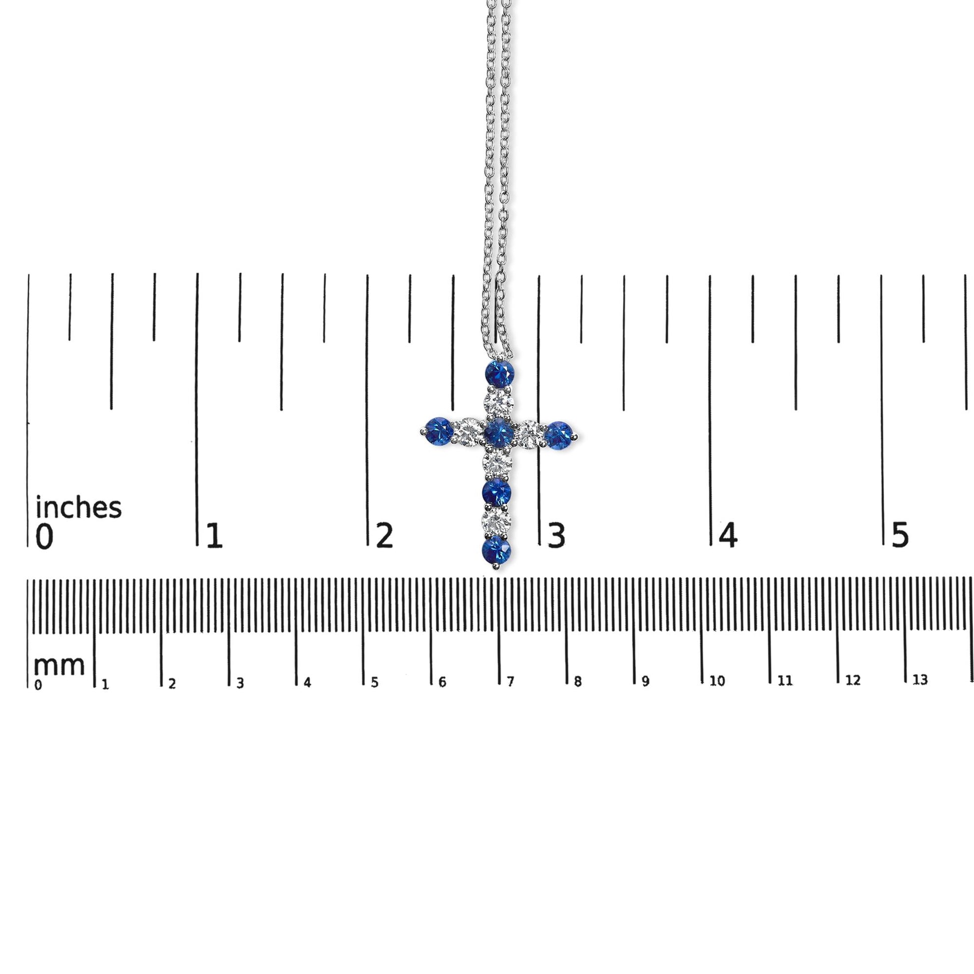White metal cross necklace with alternating round white and blue gemstones in prong settings, shown hanging vertically on a ruler for size reference viewed from the front.