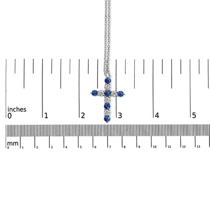 White metal cross necklace with alternating round white and blue gemstones in prong settings, shown hanging vertically on a ruler for size reference viewed from the front.