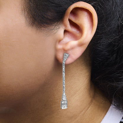 Close-up side view of a white metal drop earring on a model’s ear, featuring round bezel-set white gemstones near the earlobe and a dangling line of channel-set baguette-shaped white gemstones ending with a larger baguette-shaped gemstone at the bottom.