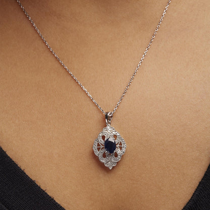 Close-up angle of a white metal necklace on a model, featuring an ornate pendant with a central oval blue gemstone surrounded by round white gemstones in shared prong settings.