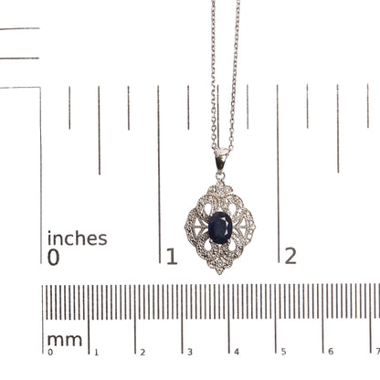 White metal necklace with an intricate pendant featuring a central oval blue gemstone surrounded by small round white gemstones, photographed from the front on a ruler for size reference.