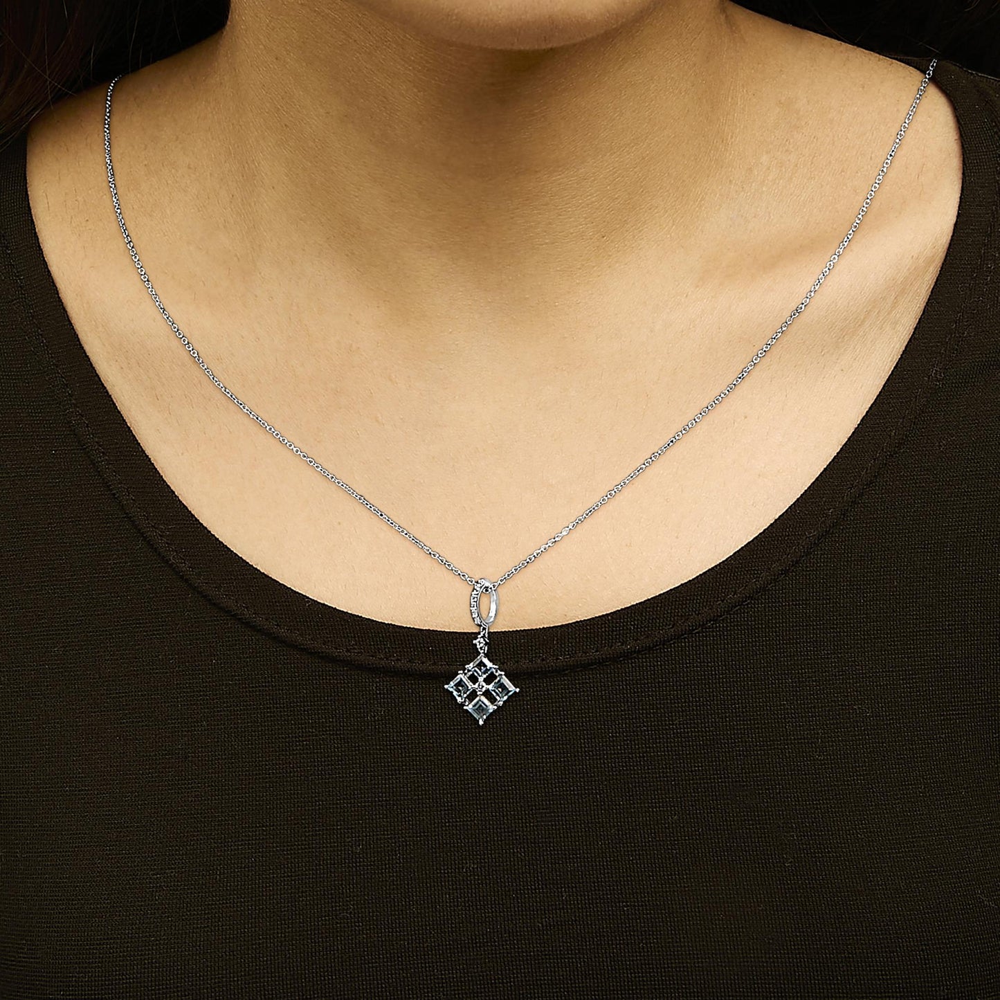 White metal necklace with a pendant featuring four square blue gemstones arranged in a diamond shape, surrounded by several small round white gemstones, shown worn on a model in a front close-up view.