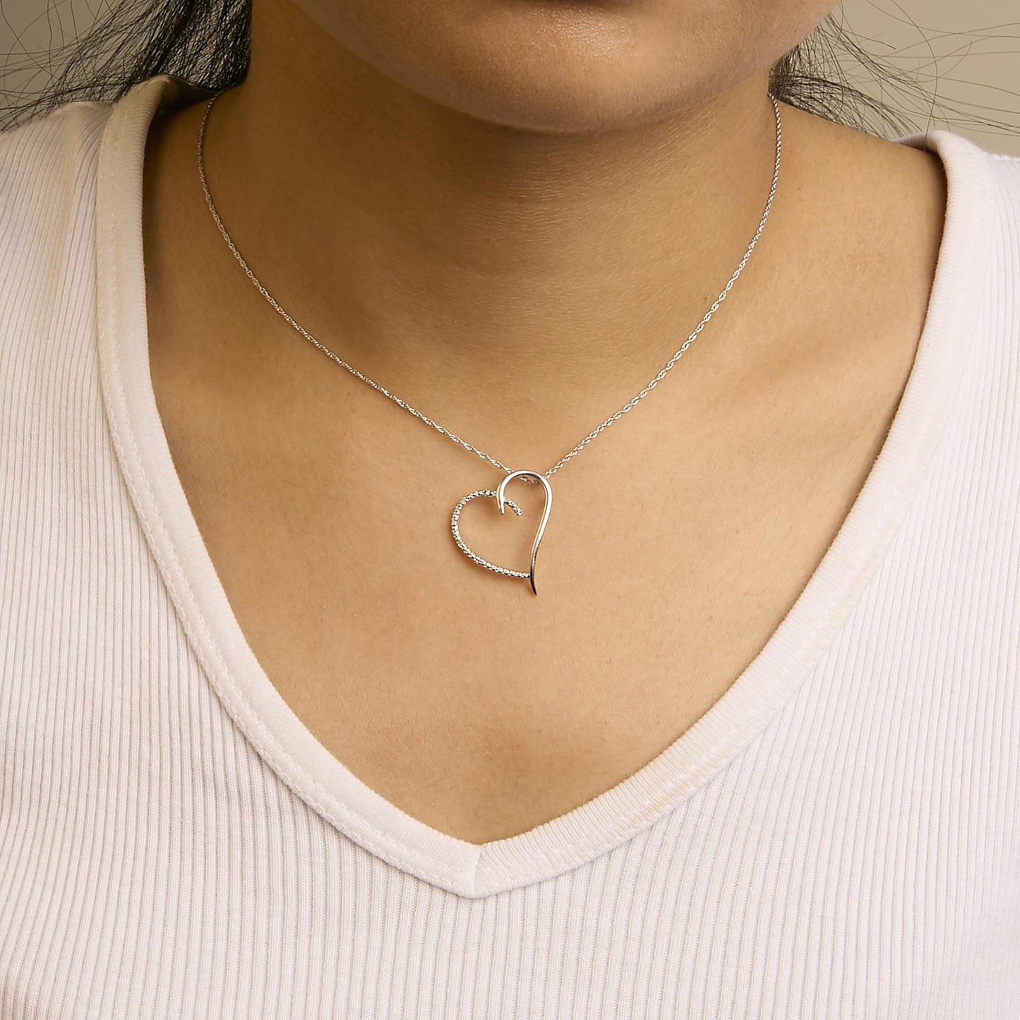 Necklace in white metal with an open heart-shaped pendant, one half plain and the other half adorned with round white gemstones, shown on a model from the front angle.