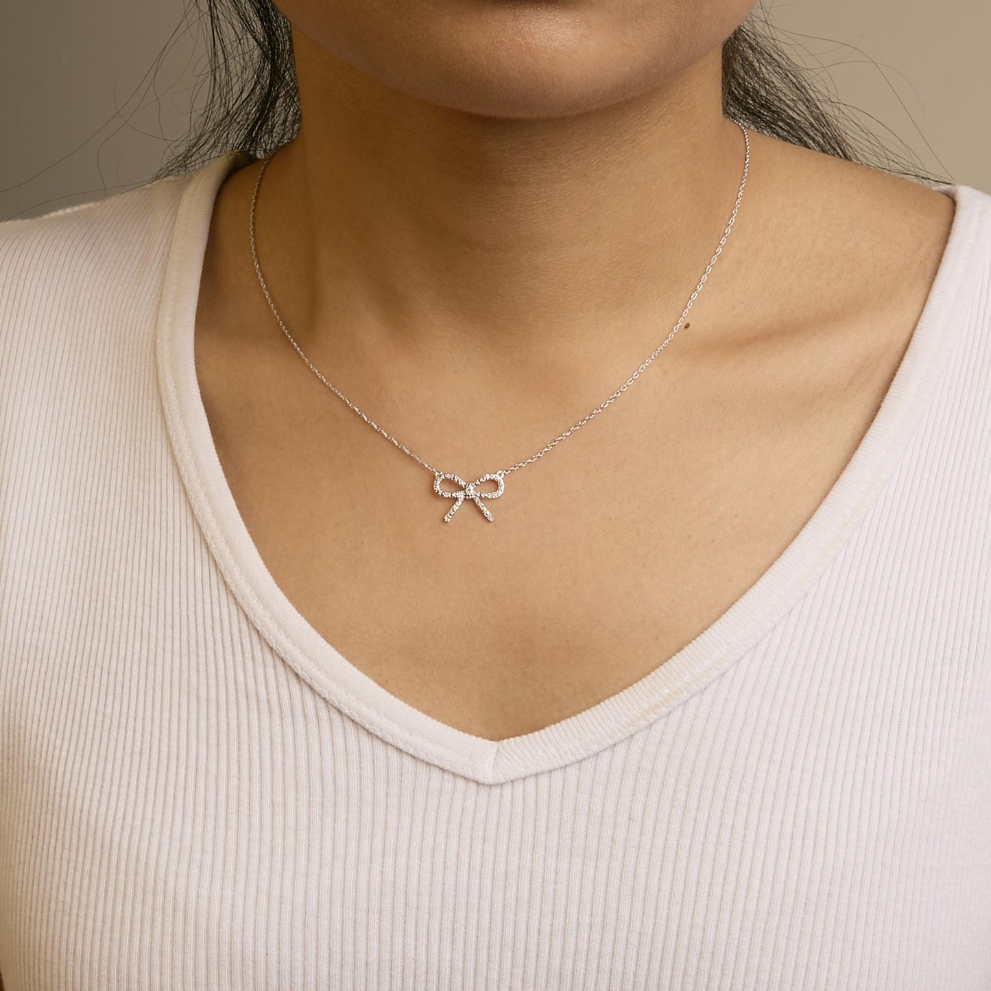 Close-up front view of a model wearing a white metal necklace with a bow-shaped pendant adorned with round white gemstones in prong settings.