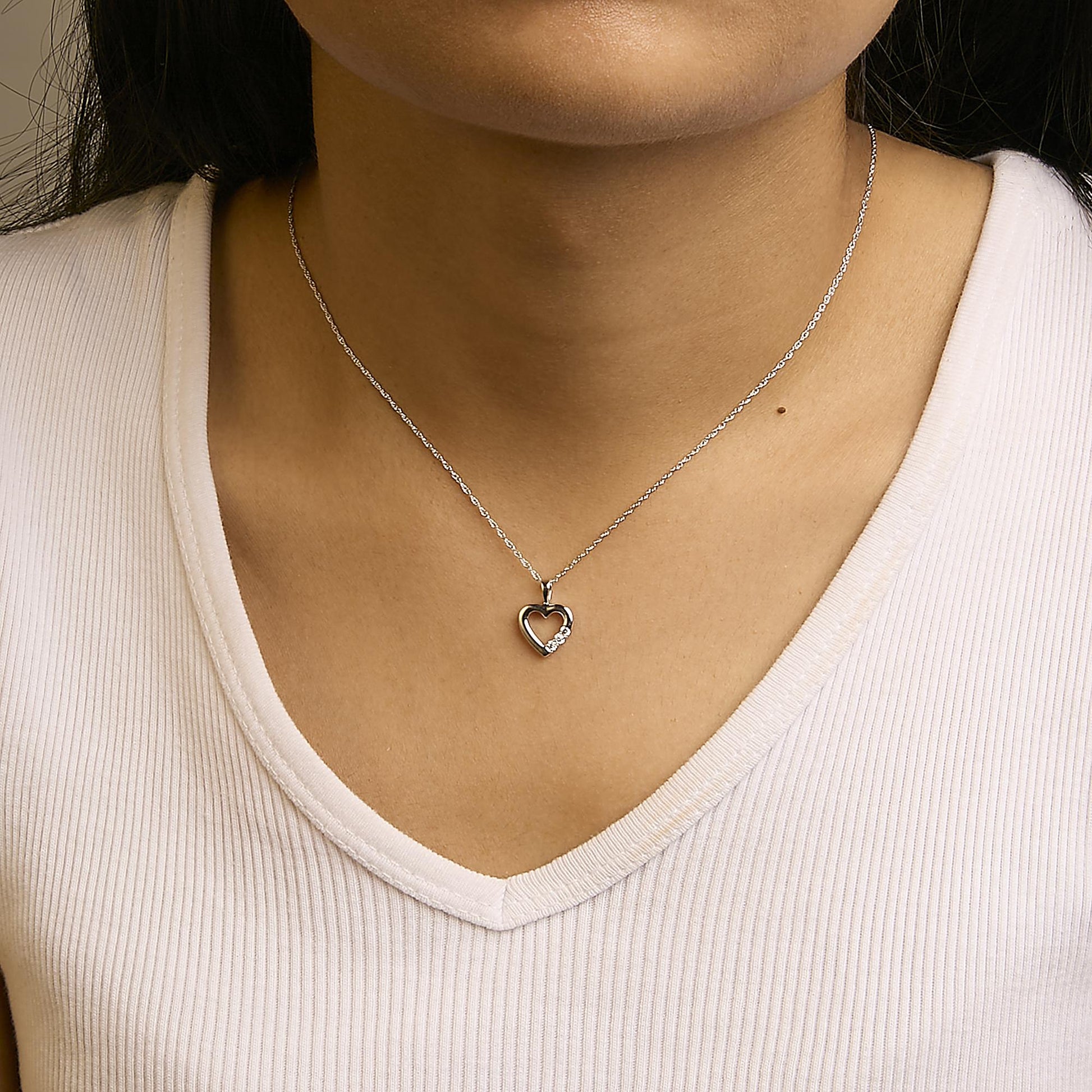 Close-up front view of a model wearing a white metal necklace with a heart-shaped pendant featuring three round white gemstones in prong settings on one side.