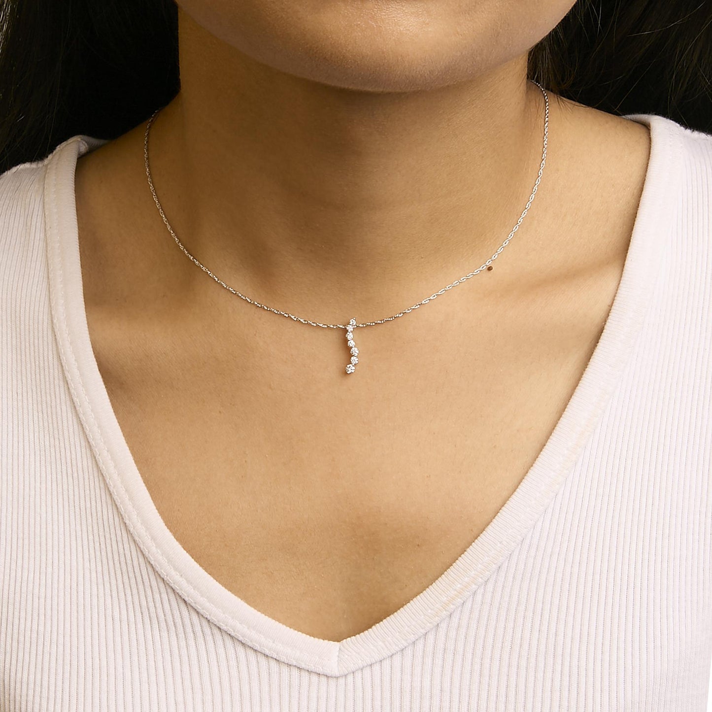 Close-up front view of a white metal necklace with a vertical pendant featuring seven round white gemstones in prong settings, worn on a model.