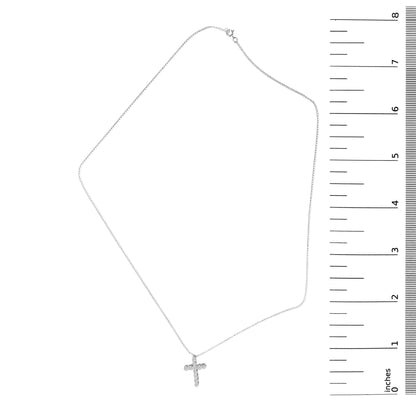 White necklace with a cross-shaped pendant featuring round white gemstones in shared prong settings, shown laid flat next to a ruler for scale, captured from a top-down angle.