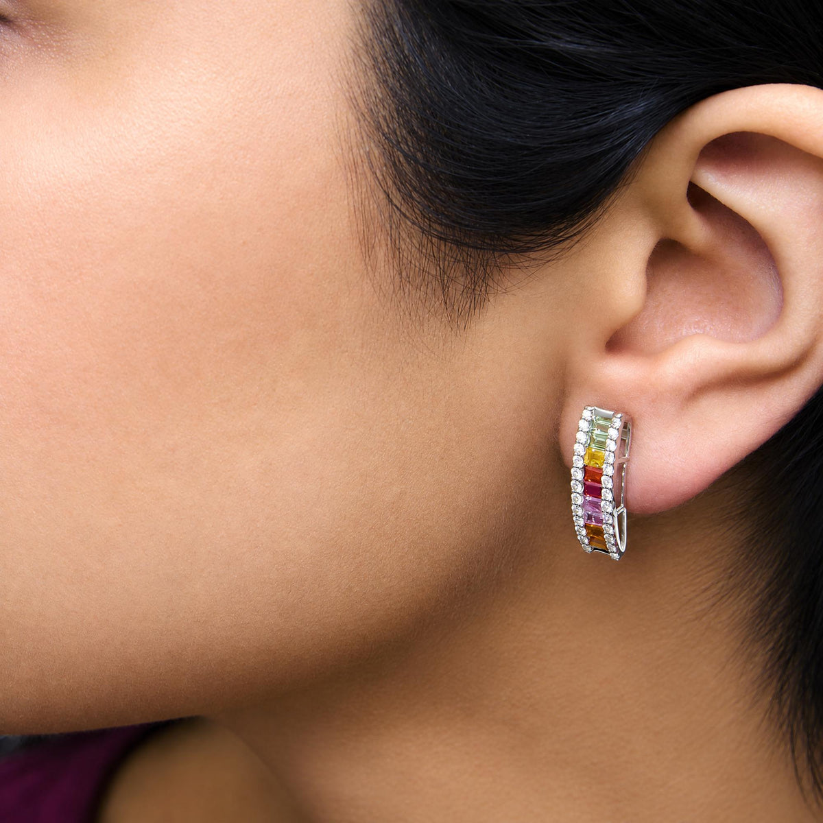 Close-up side view of a white hoop earring worn on a model's ear, featuring a row of white round gemstones in prong setting surrounding colorful baguette-shaped gemstones in channel setting in yellow, green, red, pink, and orange colors.