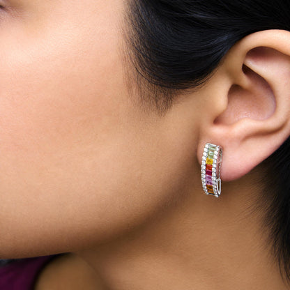 Close-up side view of a white hoop earring worn on a model's ear, featuring a row of white round gemstones in prong setting surrounding colorful baguette-shaped gemstones in channel setting in yellow, green, red, pink, and orange colors.