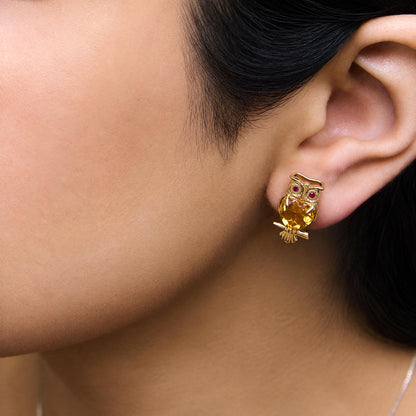 Close-up side view of a model wearing yellow metal earrings shaped like an owl, featuring two heart-shaped yellow gemstones and four round red gemstones.