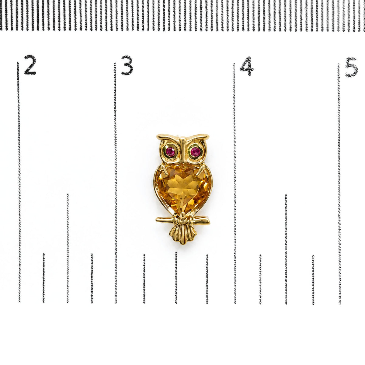 Close-up front view of yellow metal owl-shaped earring with a large heart-shaped yellow gemstone on the body and two round red gemstones for the eyes, shown on a scale.