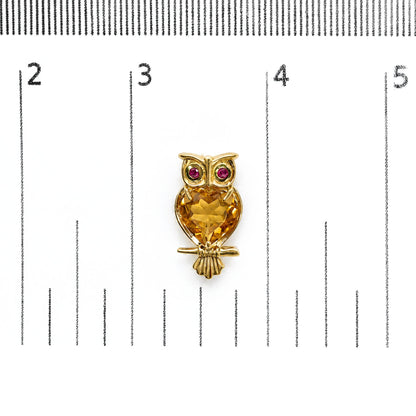 Close-up front view of yellow metal owl-shaped earring with a large heart-shaped yellow gemstone on the body and two round red gemstones for the eyes, shown on a scale.
