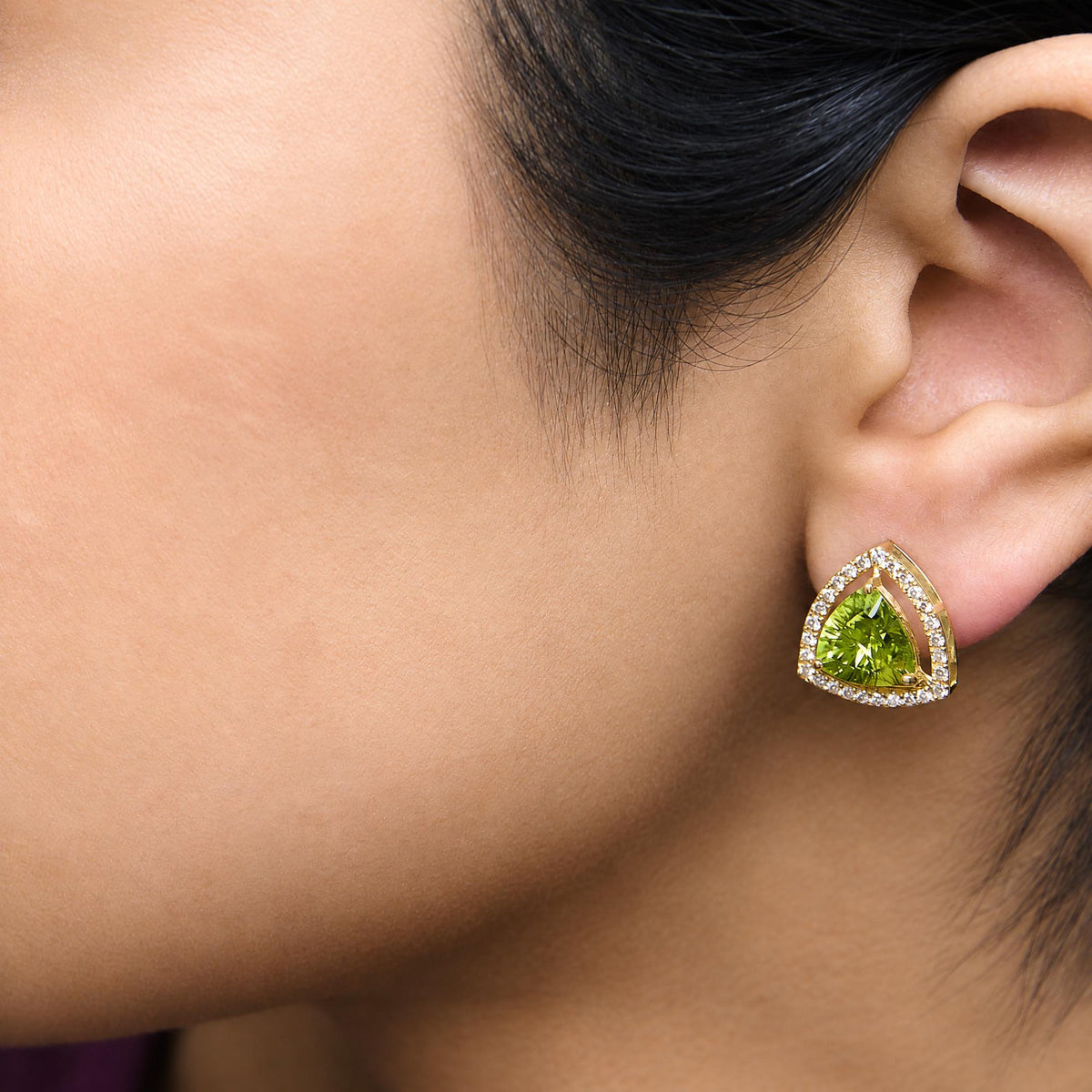 Close-up side view of yellow metal earrings shown worn on a model's ear, featuring a large green trillion-shaped gemstone surrounded by a border of small white round gemstones.