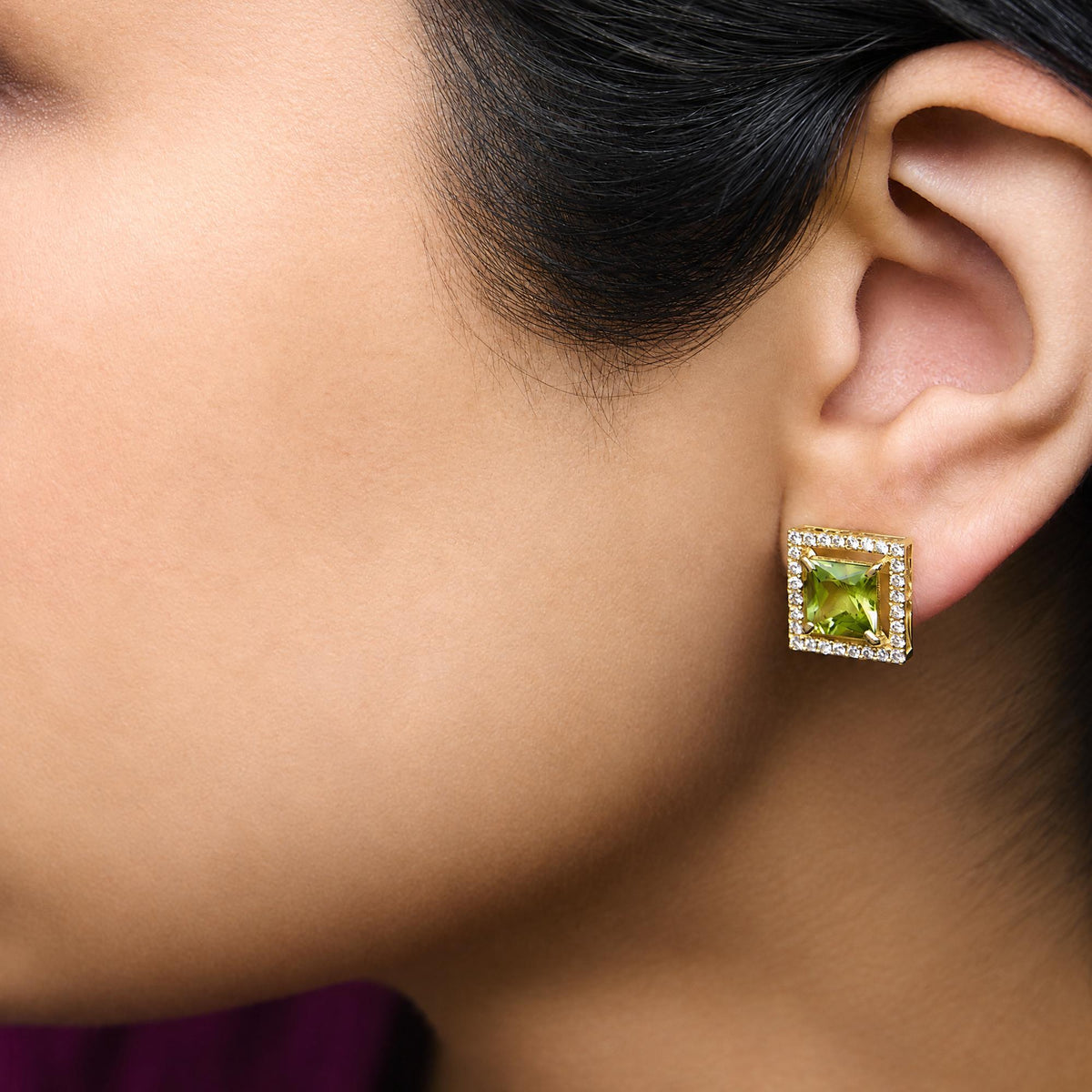 Close-up side view of a model wearing yellow metal square earrings with a large princess-cut green gemstone center, surrounded by round white gemstones in a prong setting.