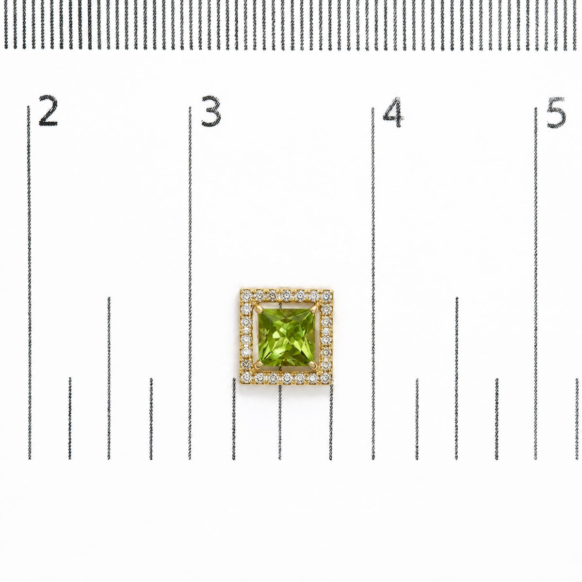 Close-up top-down view of a pair of yellow metal square earrings with a central green princess-shaped gemstone surrounded by a border of round white gemstones, shown on a scale.