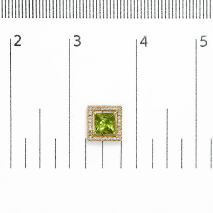 Close-up top-down view of a pair of yellow metal square earrings with a central green princess-shaped gemstone surrounded by a border of round white gemstones, shown on a scale.