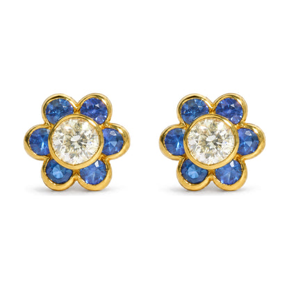 Close-up front view of pair of yellow metal flower-shaped earrings featuring a central round white gemstone surrounded by blue round gemstones in bezel settings, shown on a white background.