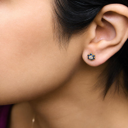 Close-up side view of a model wearing yellow metal earrings shaped like flowers with round blue gemstones and a central round white gemstone, all bezel set.