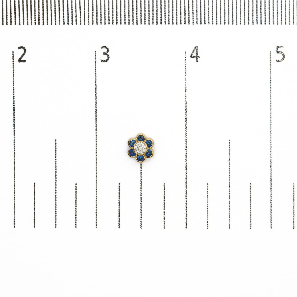 Close-up image of a yellow metal earring shaped like a flower with round white gemstone center surrounded by round blue gemstones, shown on a measurement scale from above.