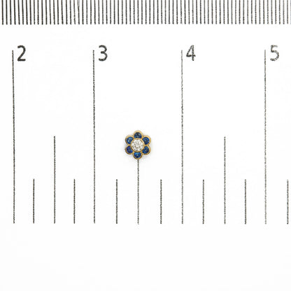 Close-up image of a yellow metal earring shaped like a flower with round white gemstone center surrounded by round blue gemstones, shown on a measurement scale from above.
