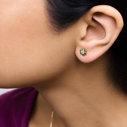 Close-up side view of a model wearing yellow metal round stud earrings with a central white gemstone surrounded by green gemstones in a floral bezel setting.