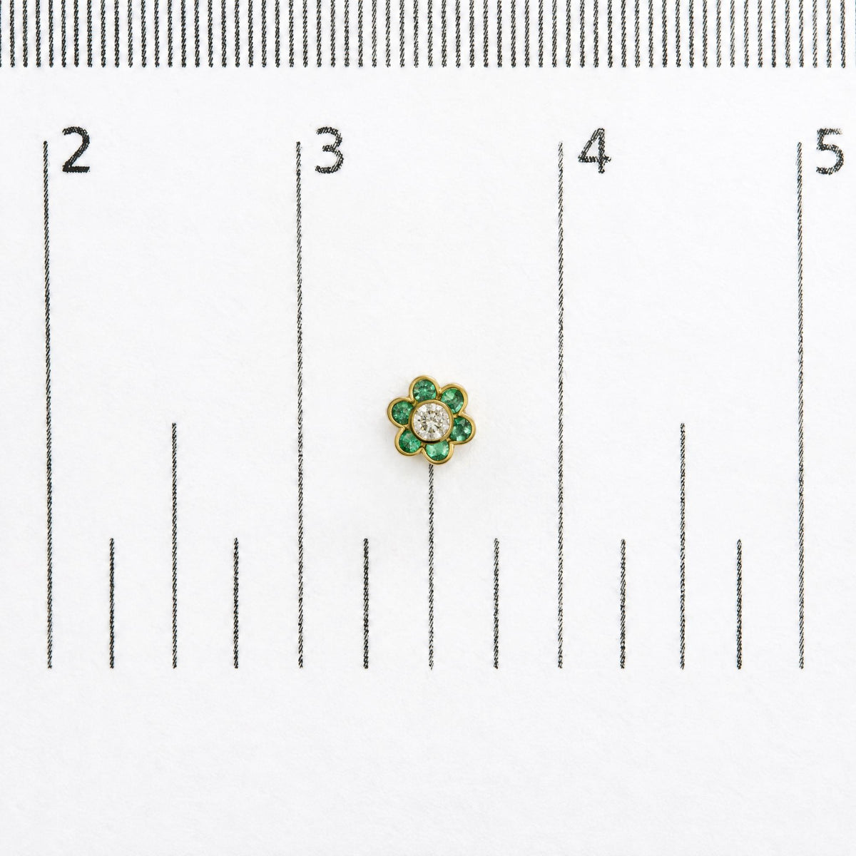 Close-up top view of a single yellow metal flower-shaped earring with round green gemstones forming petals and a round white gemstone in the center, shown on a scale background.