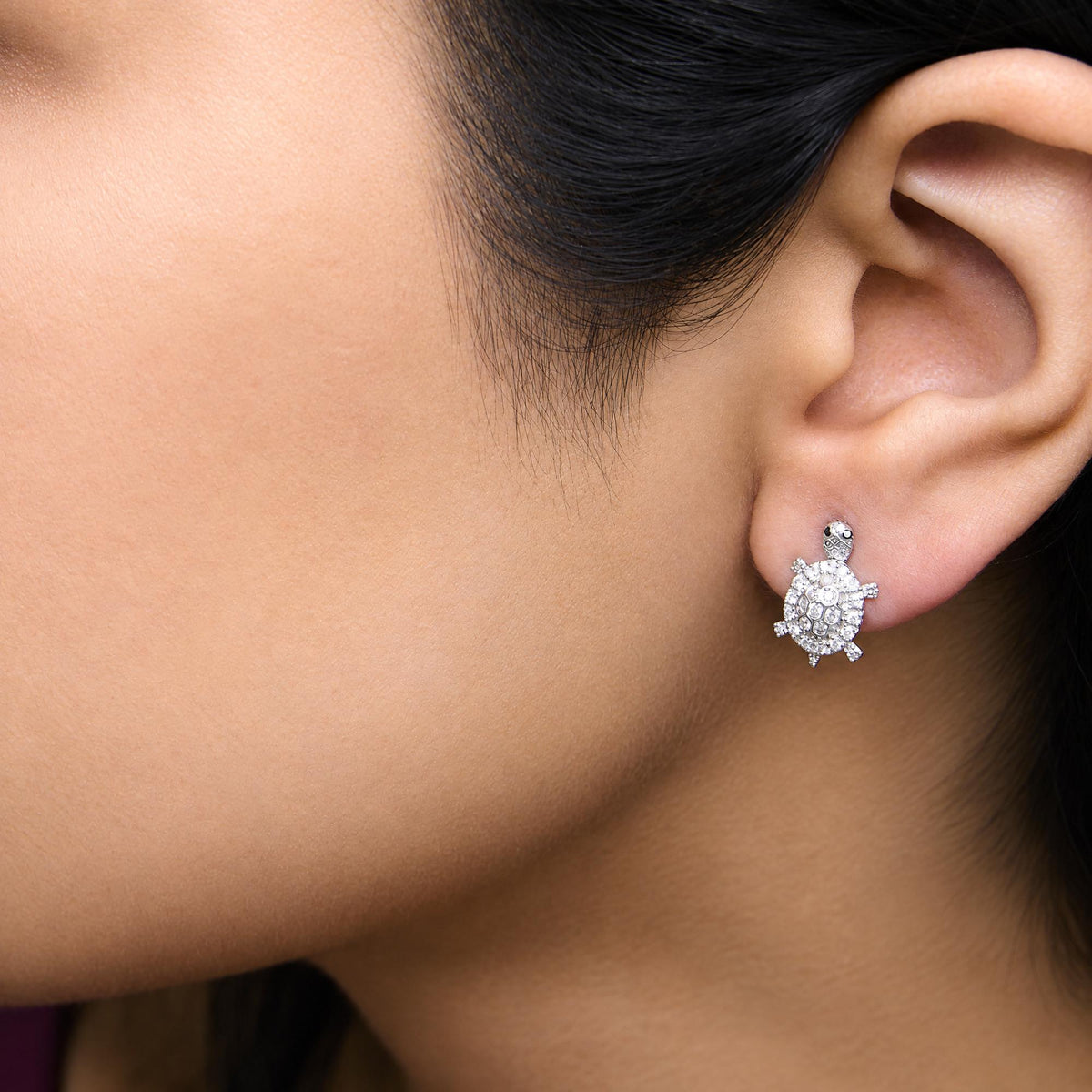 Close-up side view of a model wearing white metal turtle-shaped earrings adorned with round white gemstones in prong settings and a few round black gemstones in prong settings.