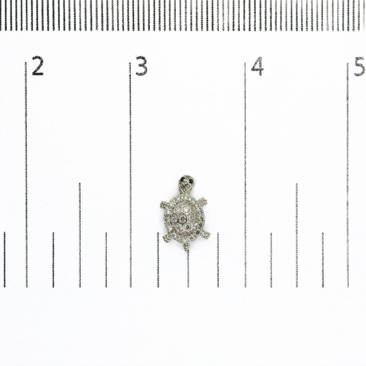 Close-up front view of a white metal turtle-shaped earring with round white gemstones and round black gemstones, shown on a measurement scale.