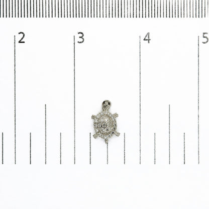 Close-up front view of a white metal turtle-shaped earring with round white gemstones and round black gemstones, shown on a measurement scale.