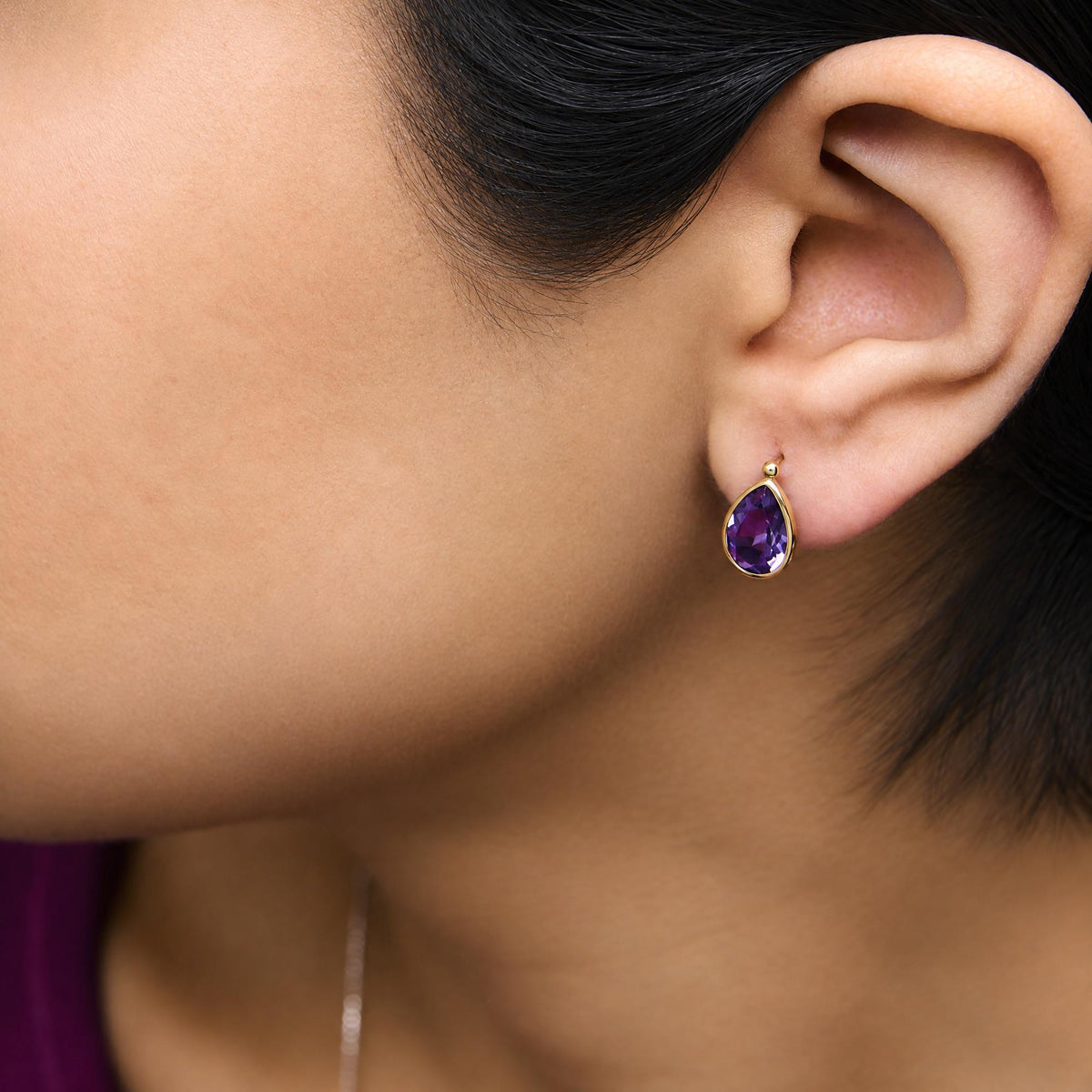 Close-up side view of a model wearing a yellow metal earring featuring a pear-shaped purple gemstone in a bezel setting.