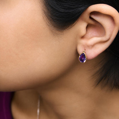 Close-up side view of a model wearing a yellow metal earring featuring a pear-shaped purple gemstone in a bezel setting.