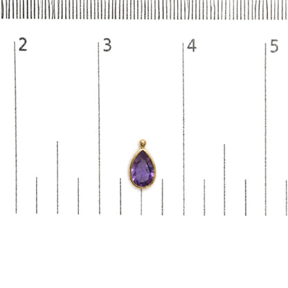 Close-up front view of a yellow metal earring with a pear-shaped purple gemstone in a bezel setting, shown on a scale.