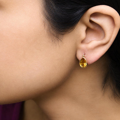 Close-up side view of a model wearing yellow metal earrings featuring pear-shaped yellow gemstones in bezel settings.