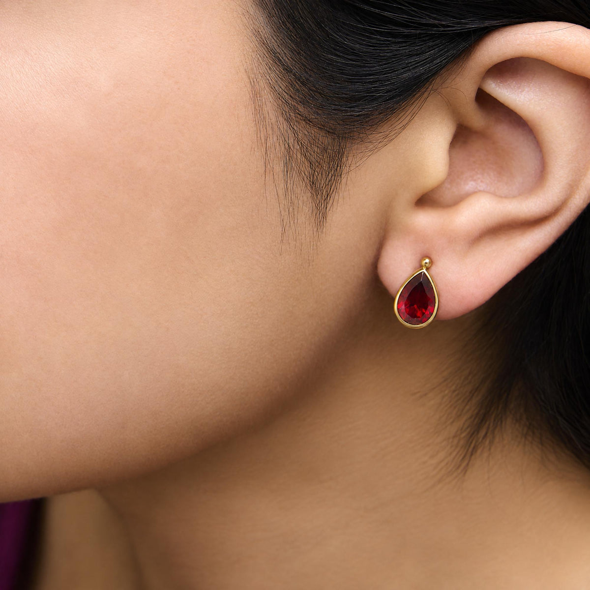 Close-up side view of a model wearing yellow metal earrings featuring pear-shaped red gemstones in bezel settings.