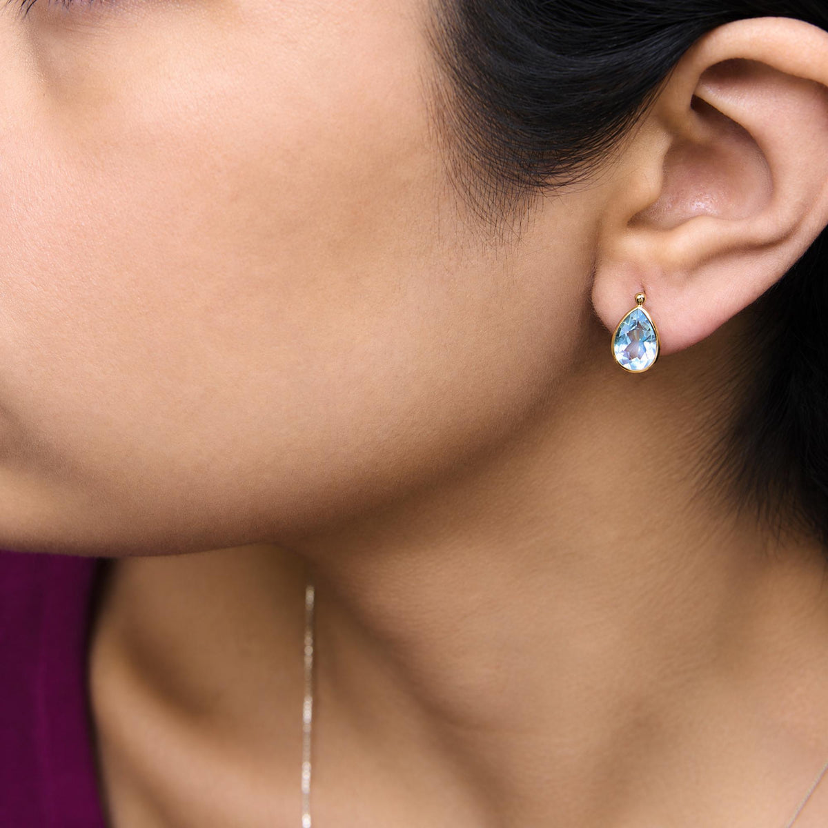 Close-up side view of a model wearing a yellow metal earring with a pear-shaped blue gemstone in bezel setting.