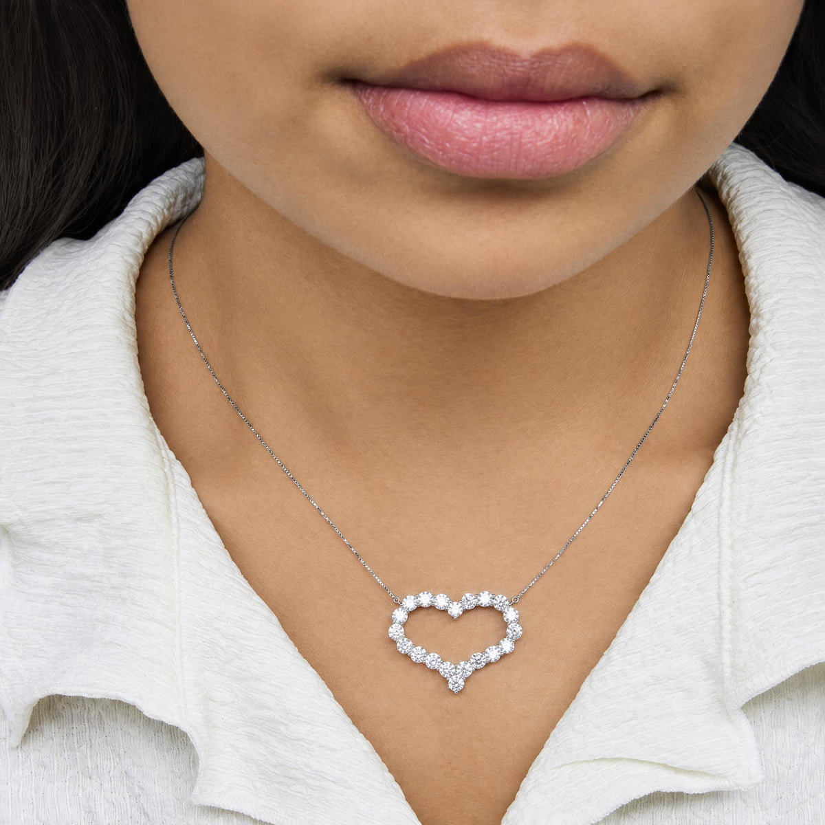 Delicate white metal necklace with a heart-shaped pendant made of round white gemstones in prong settings, shown worn on a model from a front close-up angle.