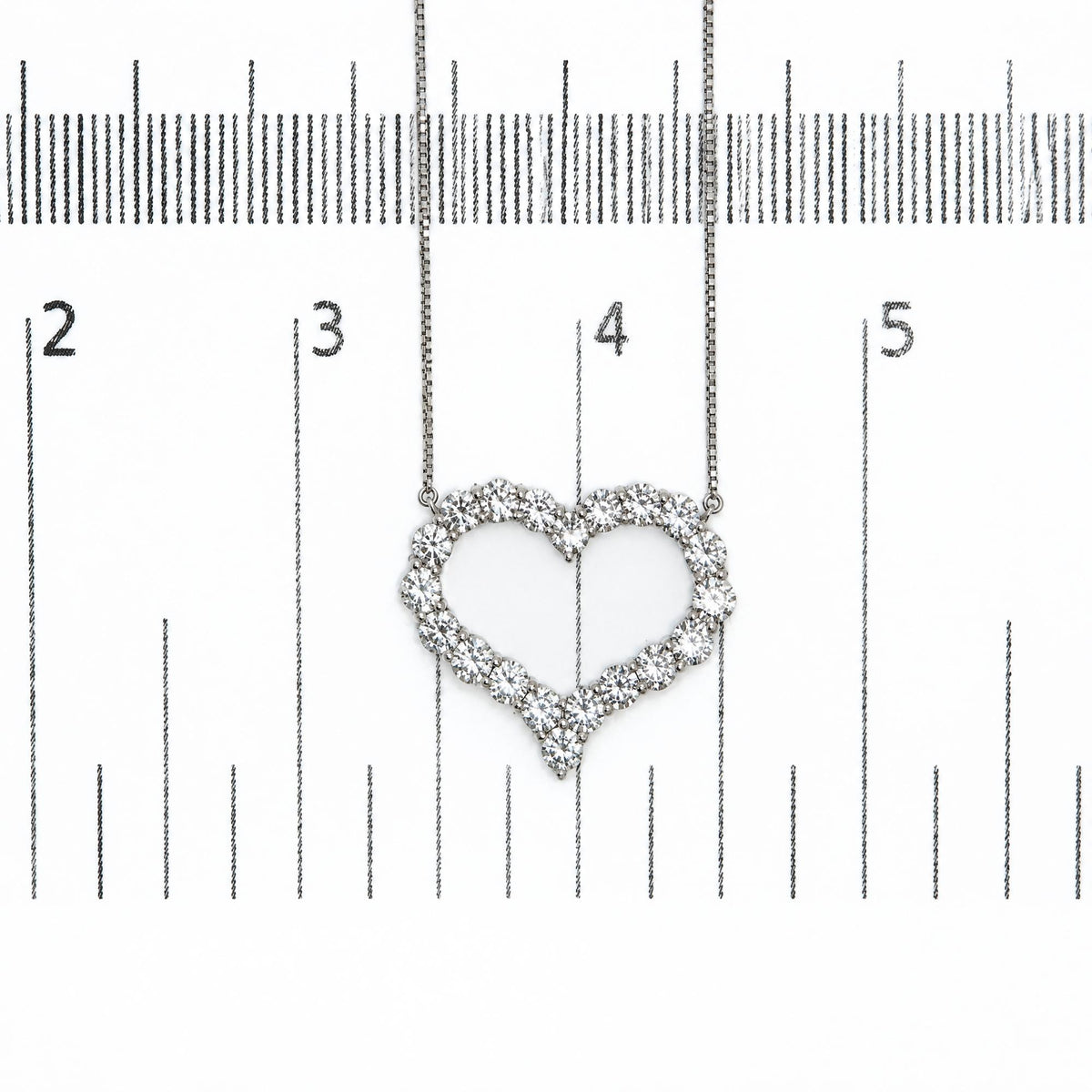 Close-up of a heart-shaped necklace pendant with white round prong-set gemstones, shown on a ruler scale from a top-down angle.